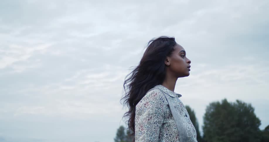 Woman walks under cloudy sky near trees as wind blows through her hair