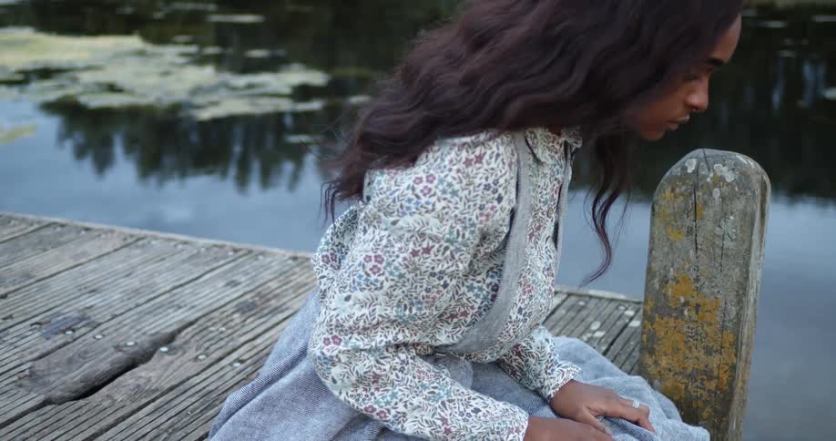 A woman sitting on the edge of a wooden dock lifts her skirt as she looks down into the water