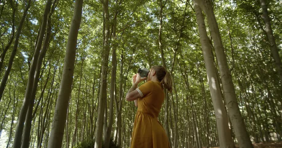 Young woman in a yellow mustard dress filming the trees on a forest at day time