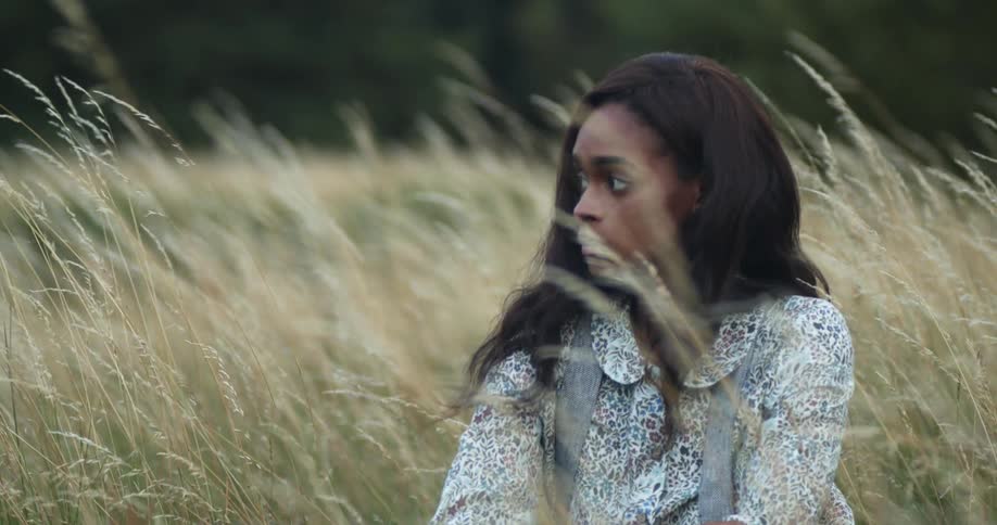 A young woman sits contemplatively in the field of tall grass during the day