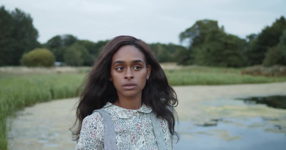 A black woman posing in casual clothing by a pond near a forest