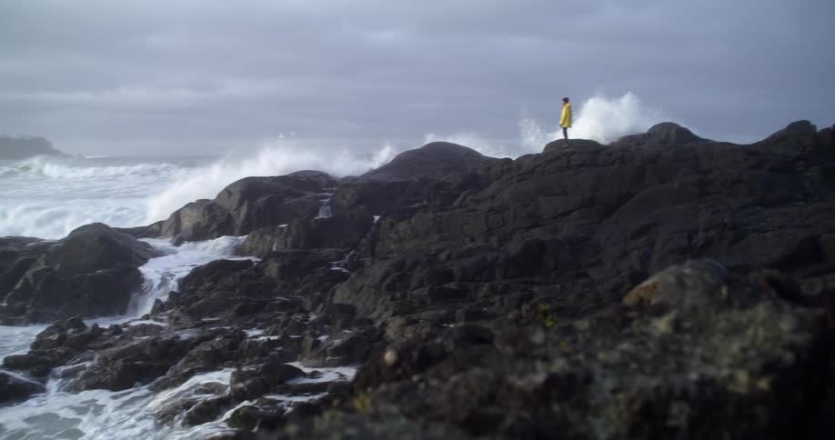 Person wearing yellow jacket is standing on rocky coastal bank as waves crash against the rocks and splash high on a cloudy day