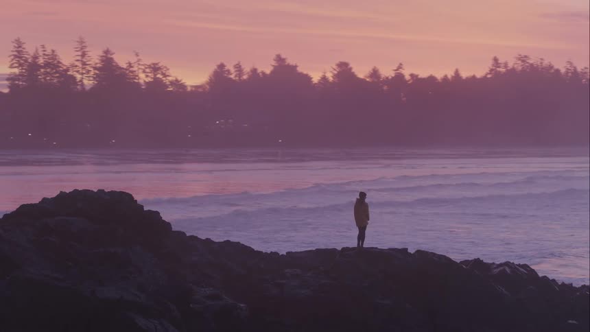Aerial of a woman standing on a rocky outcrop watching the waves at sunrise
