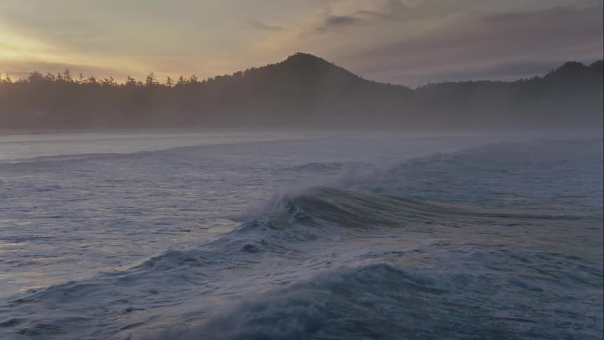 Aerial view of the mist floating over the ocean waves next to the mountains at sunrise