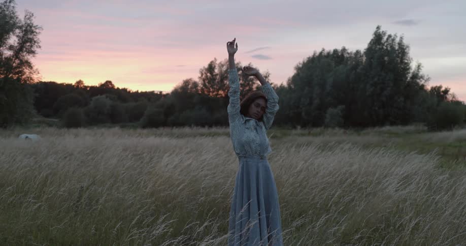 A model in a prairie dress poses with her arms above her head while standing in a field with tall weeds