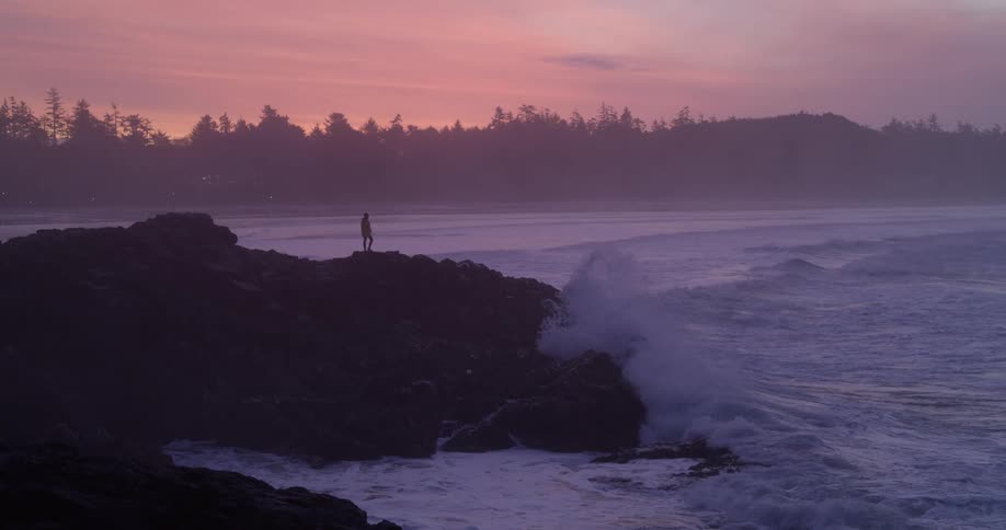 Person standing overlooking the ocean at the edge of a small sea cliff as waves crash up on the sides in the evening