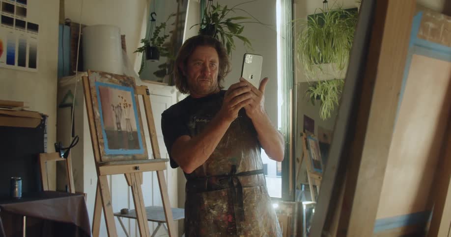 Bearded painter takes photos of his pictures using a smartphone in a studio