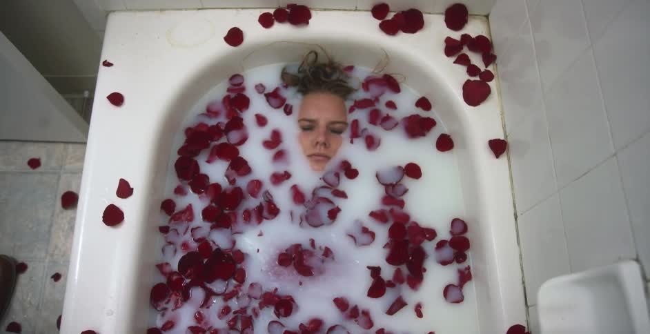 Woman in a red dress submerges in the white liquid filling the batthub with rose petals floating