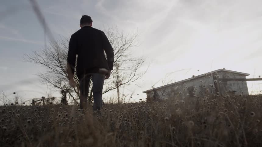 A man walks in a weed covered field toward a farmhouse