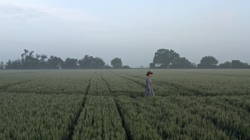 Woman wearing a hat and dress walks through field of crops with many trees in the distance