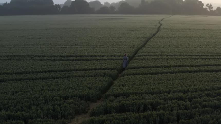 A woman walks on a path in between small shrubs in a wide open field in the evening