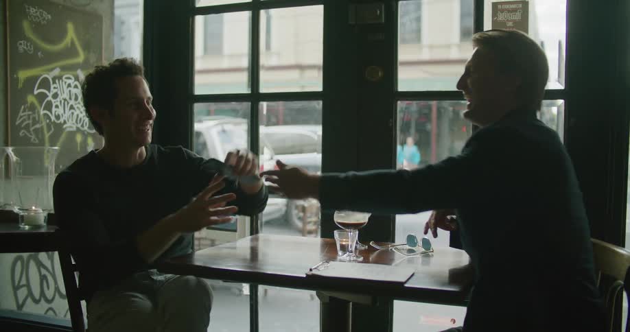 Two men sitting at a table in a restaurant enjoying a drink looks at a cell phone as vehicles drive past outside