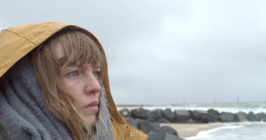 A woman stands on the beach in a yellow parka with the hood up as she stares into the distance