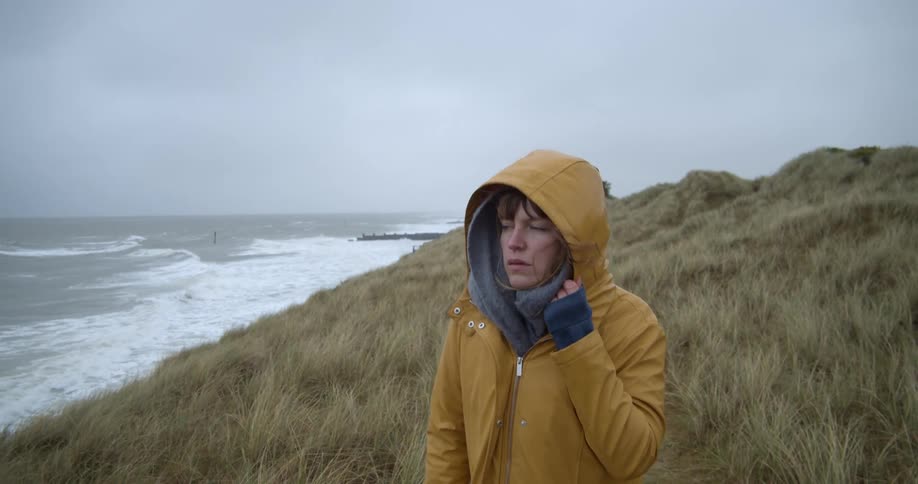 A woman in a yellow raincoat holds onto her hood while walking on a hill along the coast of the sea