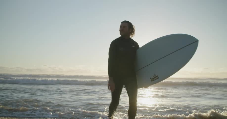 Man walks out from water in the ocean in front of a flare of sunlight holding a surfboard