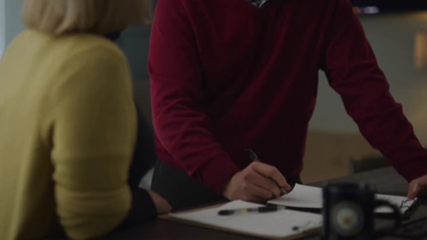 A man standing in front of a desk in an office writes on a clipboard lying on the tabletop while speaking to a woman standing next to him