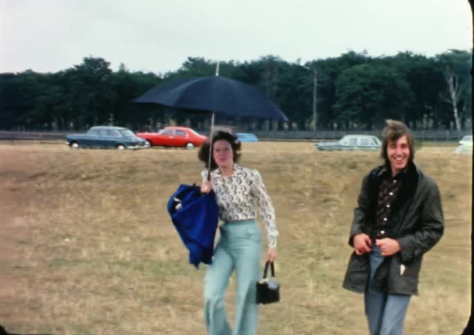 A woman holds a black umbrella over her head and walks next to a man in a grass field smiling at the camera during the day