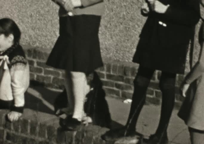 Black and white footage of a young boy who runs over to a group of children and hides behind a girl