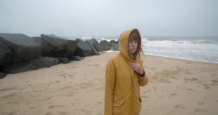 A woman holds her yellow rain jacket closed as she stands on the beach