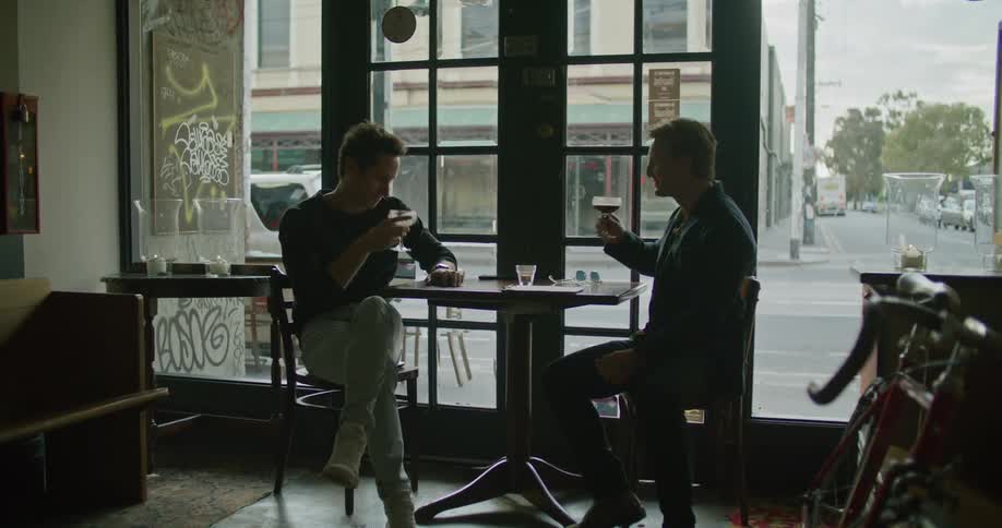 Yellow colored train passing behind two men drinking and toasting in a small bar