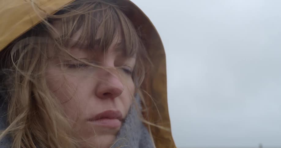 A woman in a hooded jacket stands outdoors looking pensively into the distance