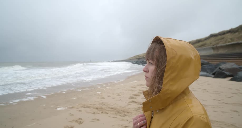 A woman holds her yellow raincoat closed with her hand as she walks on the beach