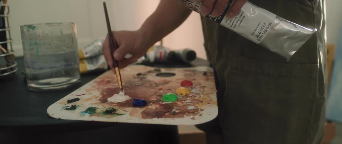 A teenage asian girl places white paint on a board with a paint brush as she stands indoors
