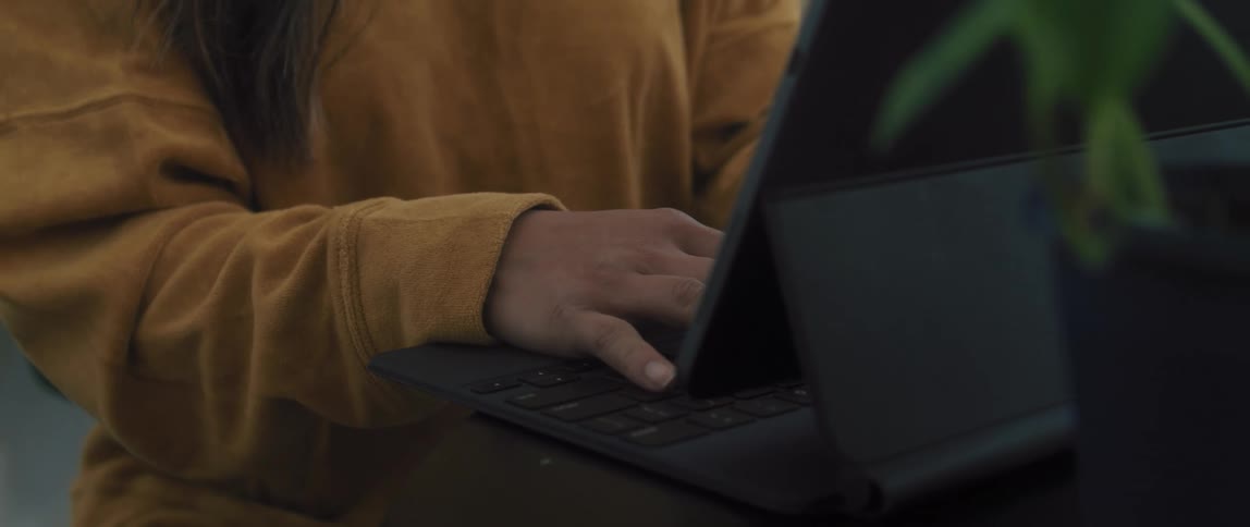A woman with a mask sitting on a chair at a table typing on a tablet