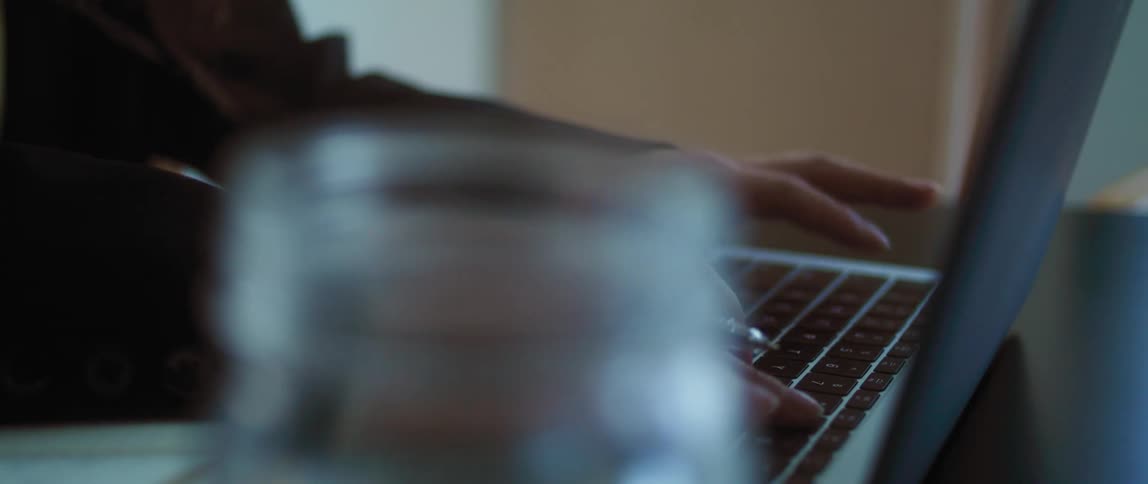 A person typing on a laptop with a pen in their hand while sitting at a desk