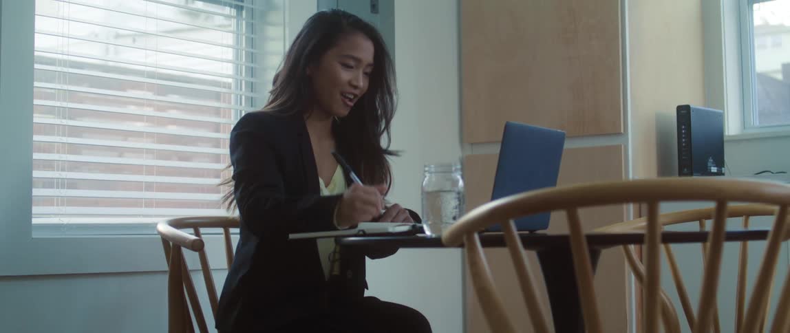 A young woman in business attire sitting in a dining room video chatting on her laptop