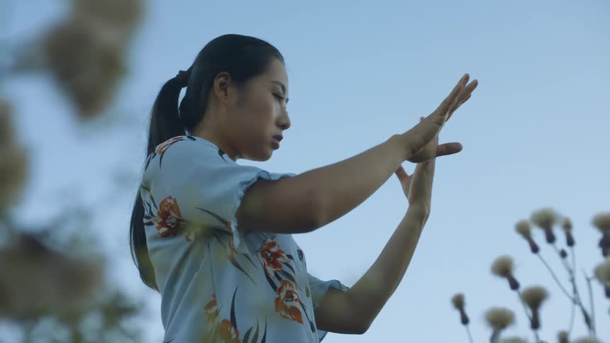 A woman stands in a field of flowers and looks at her hands and palms in confusion