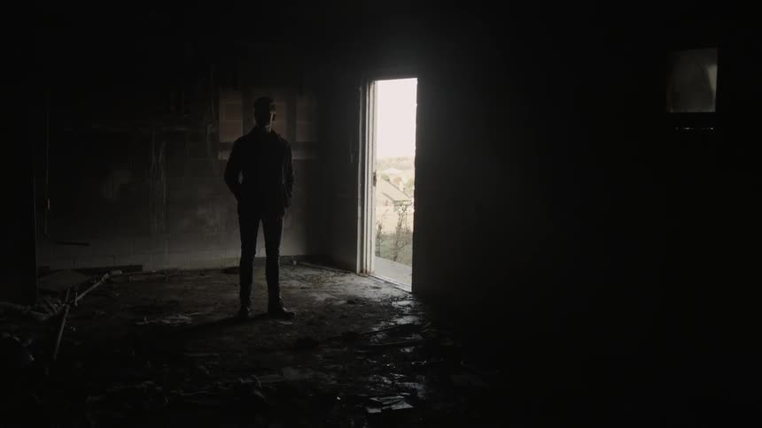 Man standing still in an empty dark room near an open door