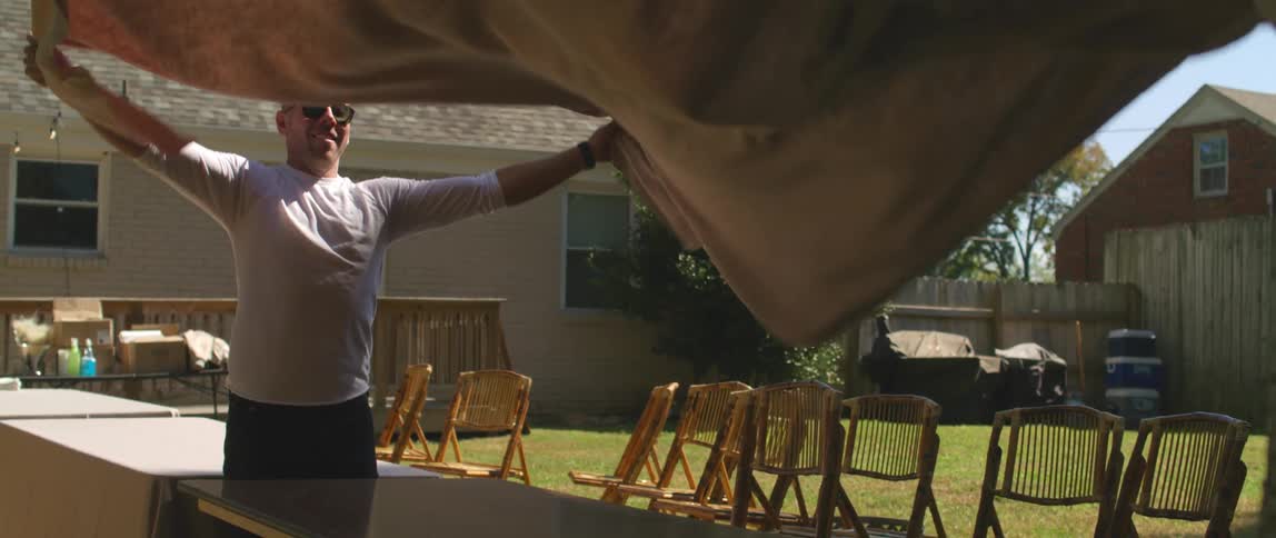A man smiles as he and a partner shake out and spread a tablecloth in the backyard