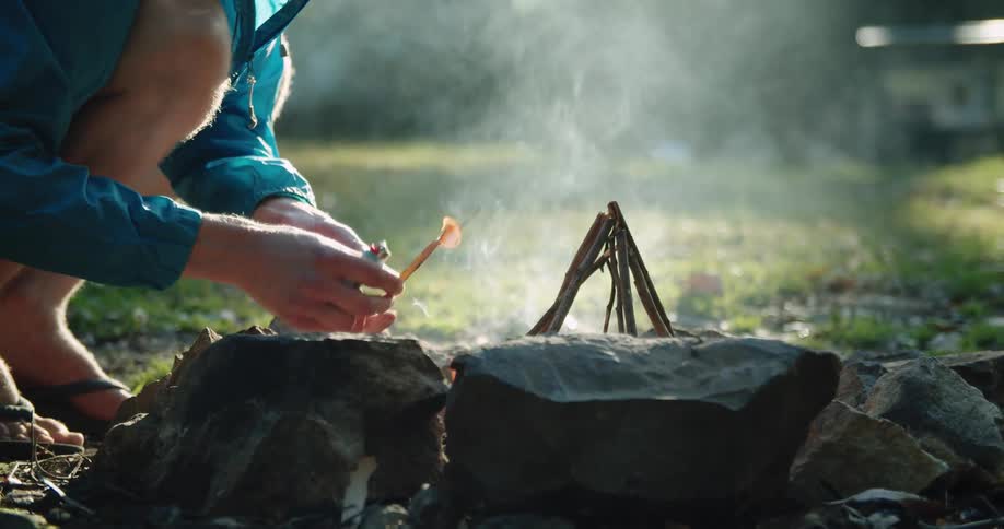 A man knees down and blows on kindling as he tries to light a fire while camping
