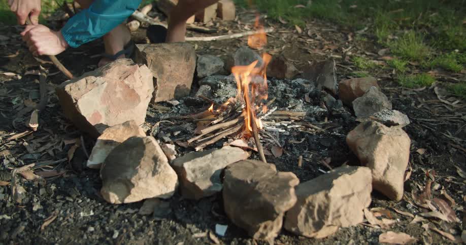 A person sitting on a camping chair beside a campfire uses a stick to shift a stone of the fire circle