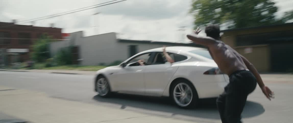 A car full of people in a white sedan wave at a teenager rolling skating down a city side street