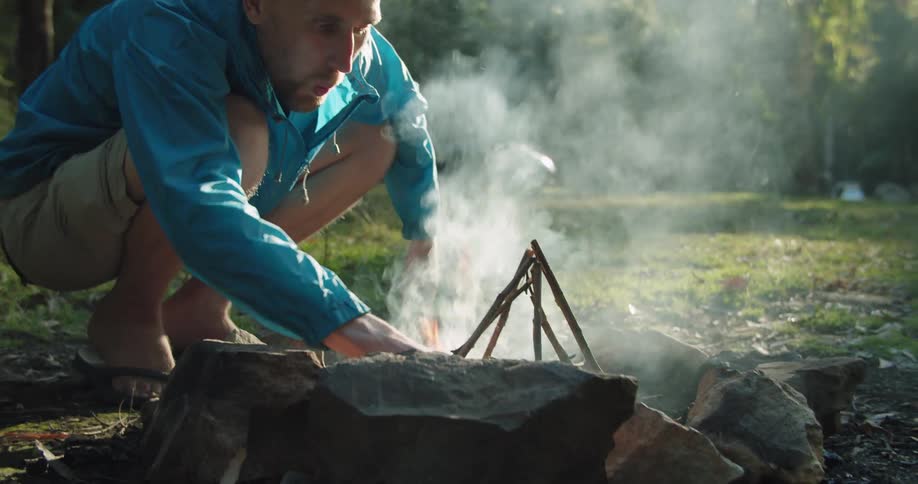 Man kneeling in front of a campfire starting a fire near the woods at morning