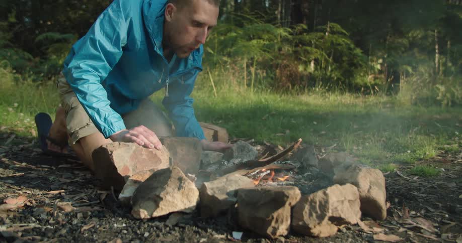 A man kneels on the ground in the forest and blows out a campfire