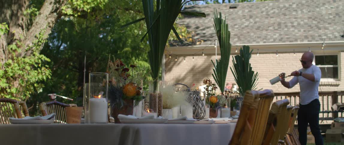 Bald young man light candles in the center of dining tables on the backyard at evening