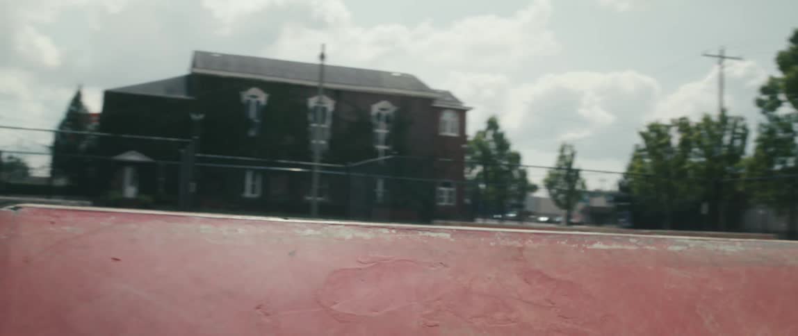 A man in a skate park doing tricks with in line skates with trees buildings sky and clouds in the background