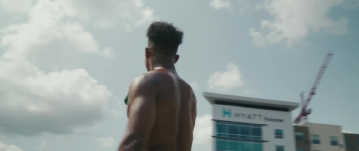 A male stands outside with no shirt and drinks from a soda bottle then motions to someone close by