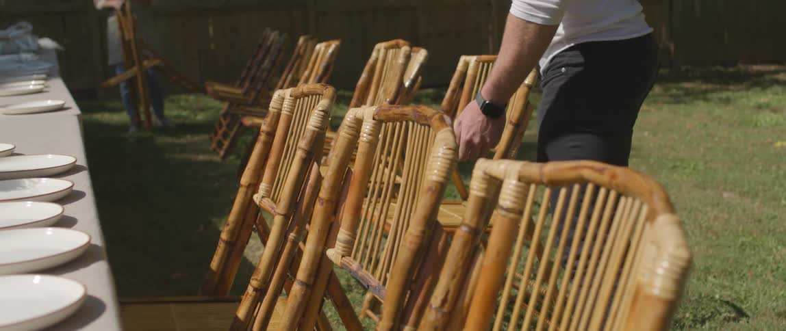 A man and woman set chairs up around a long table in a back yard