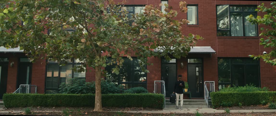 A man carrying a suitcase walks out of his apartment building and checks his phone and then smiles as his friend pulls up in a car