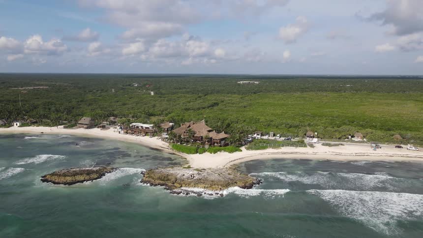 Buildings and a road on a sandy beach on the coast of a landmass covered in thick vegetation on a sunny day