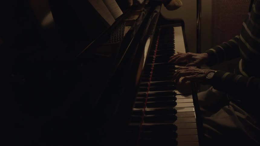 An older woman plays a piano in a dimly lit room