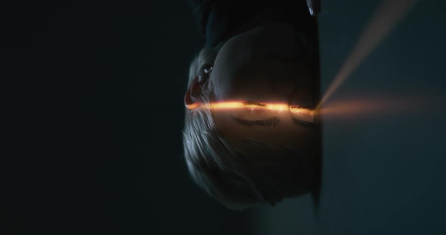 A woman lies on a cement floor with her face to one side with a beam of light shining on her eyes