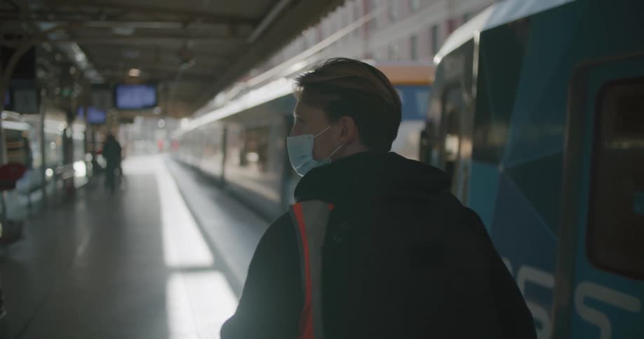 Man walks under roof of transit station past stopped public transportation wearing face mask