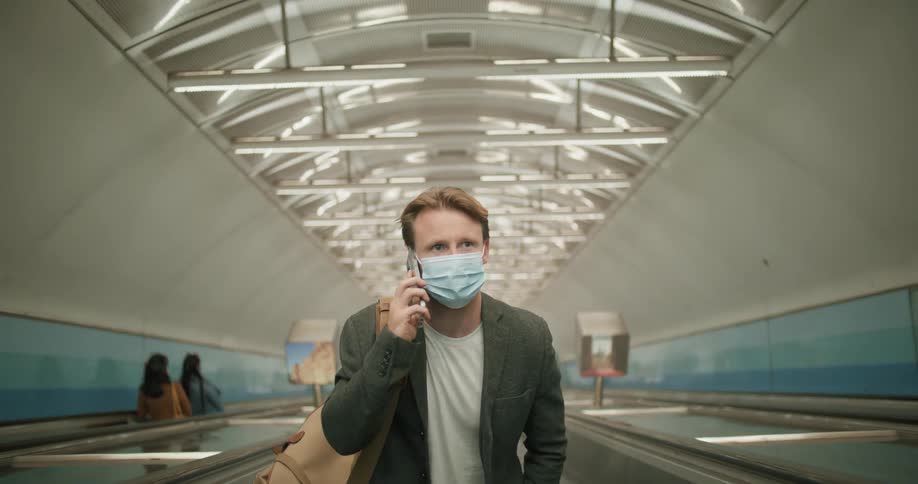 Man wearing a face mask riding an escalator while talking on a smartphone