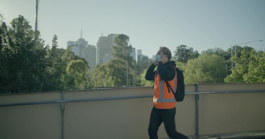 A man with a backpack walking and putting on a mask with trees buildings the sky and clouds in the background