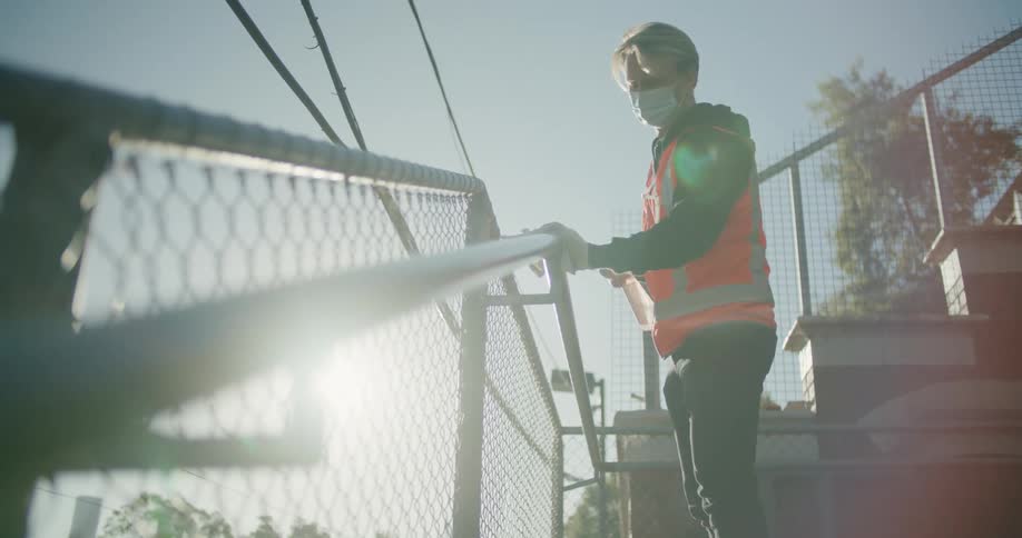 Man wearing safety vest and face mask cleans banister with spray on clear sunny day